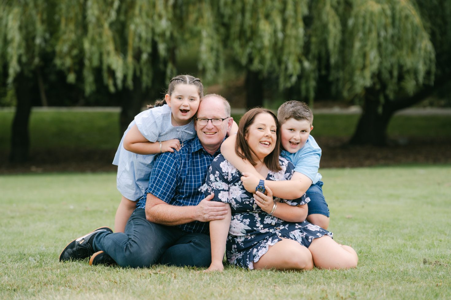 Joyful outdoor family photography session featuring parents and children laughing under a weeping willow tree.