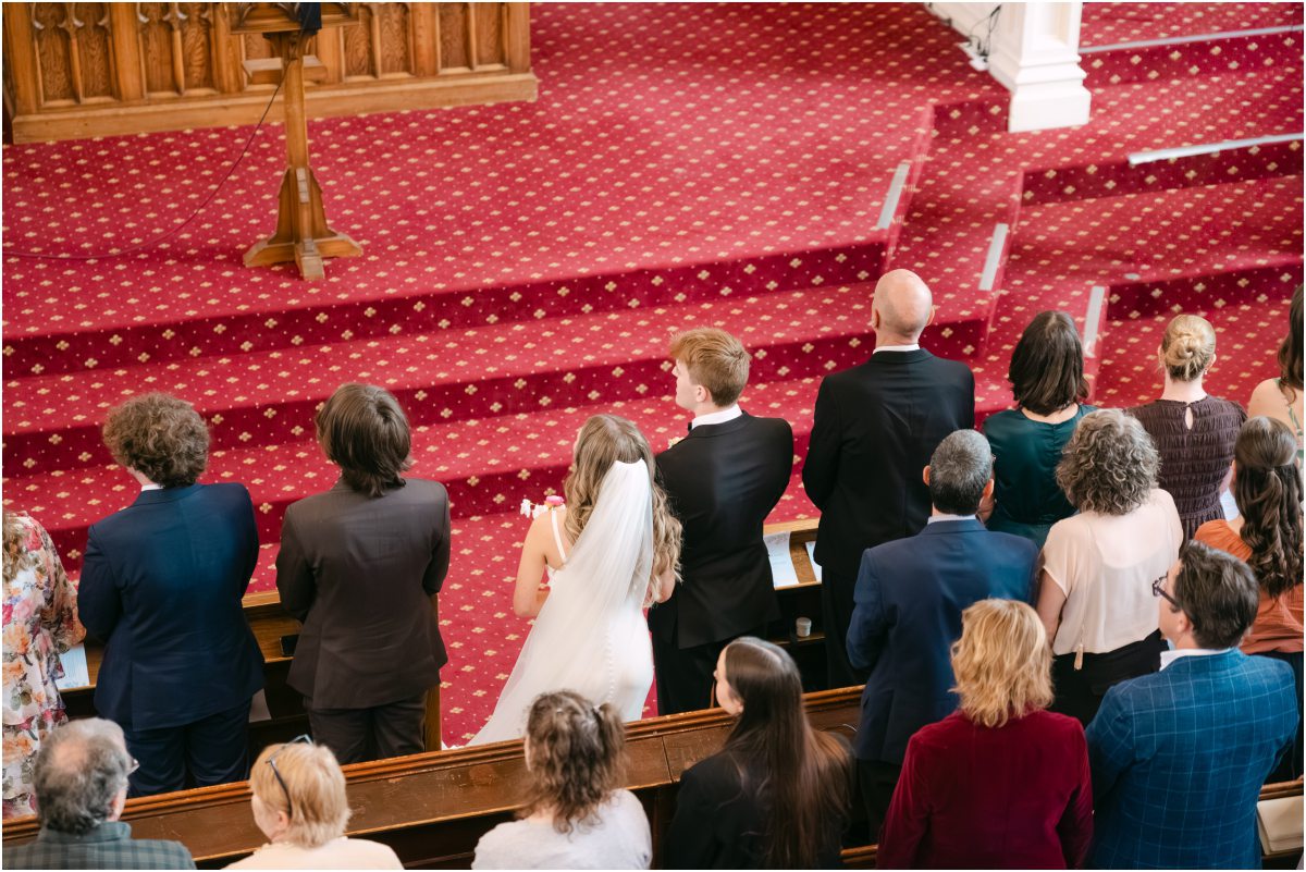 High-angle wedding ceremony photography: Bride and groom standing at the altar before the red patterned carpet steps at First Church Dunedin.