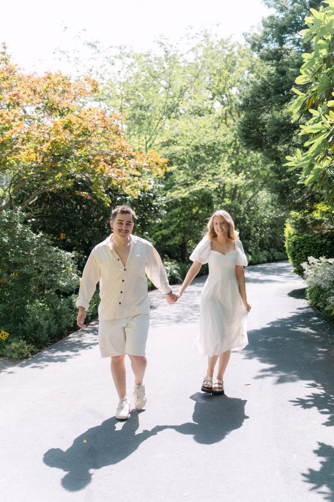 Christchurch engagement photography: Couple wearing elegant all-white outfits holding hands while walking down a sunlit garden pathway.
