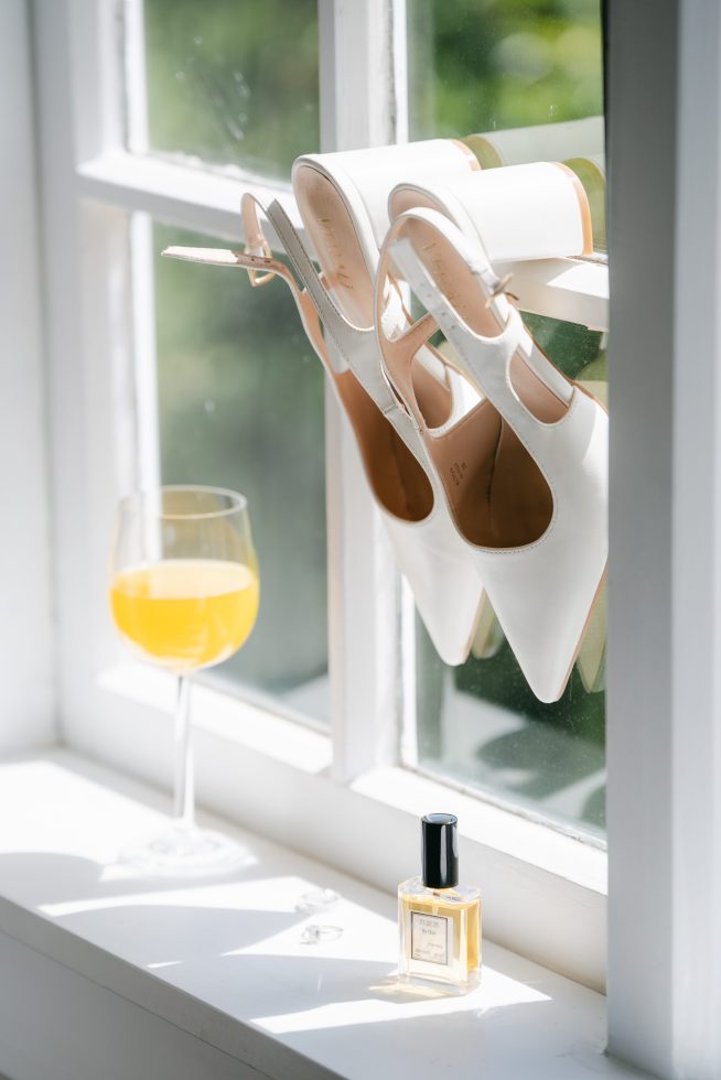 Creative bridal detail photography: White wedding heels hanging from a glass window pane next to a mimosa drink by Agnes Grace Photo & Film.