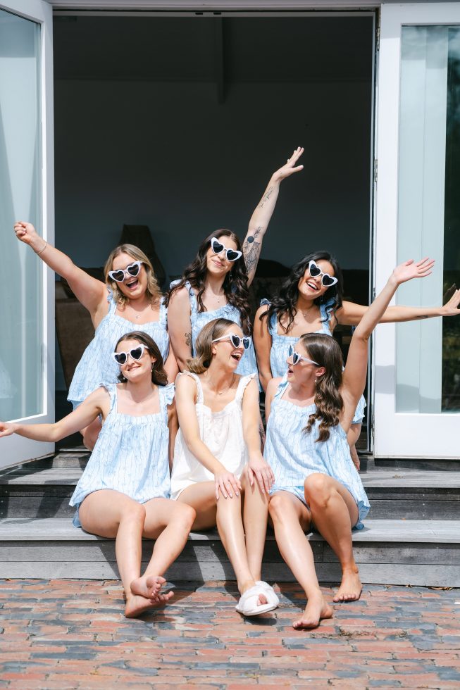 Fun bridesmaid photography: Bridesmaids in matching white pajamas and sunglasses cheering outside an estate doorway.
