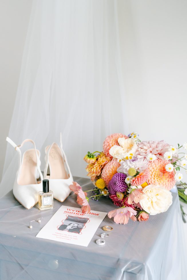 Bridal detail styling: Elegant white wedding shoes set against a sheer veil and vibrant dahlia flower bouquet.