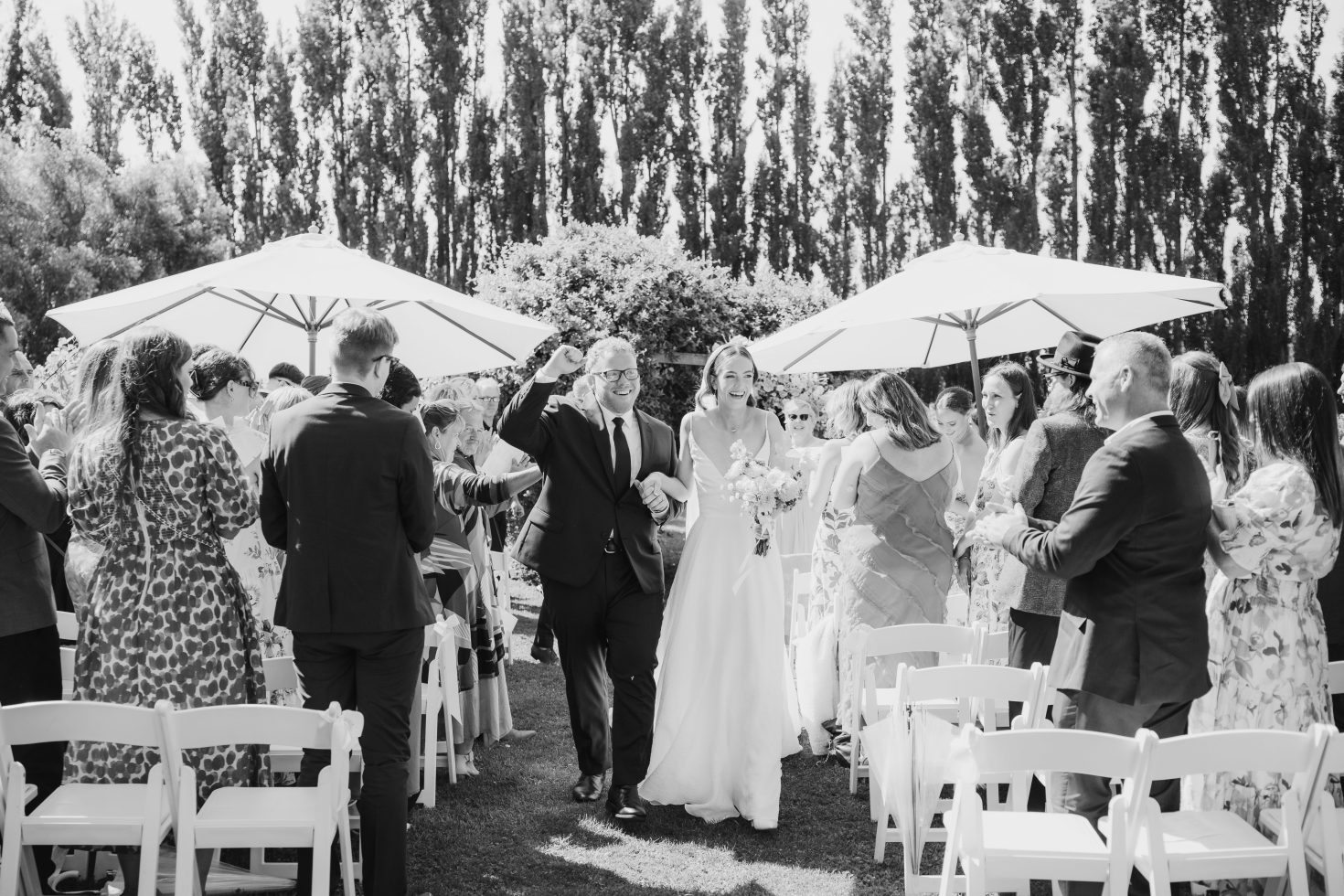 Black and white wedding photography: Blair and Ben walking down the outdoor ceremony aisle at Cossars Wineshed as guests toss flower petal confetti.
