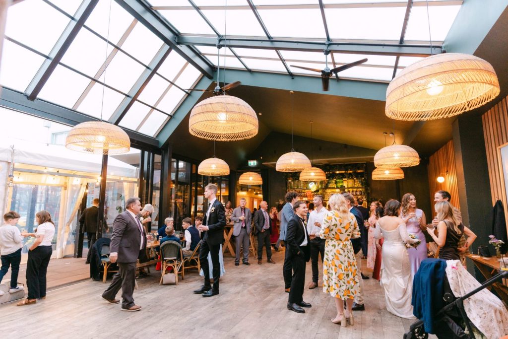 Wedding guests socializing under the warm woven basket pendant lights inside the spacious Welles & Good venue.