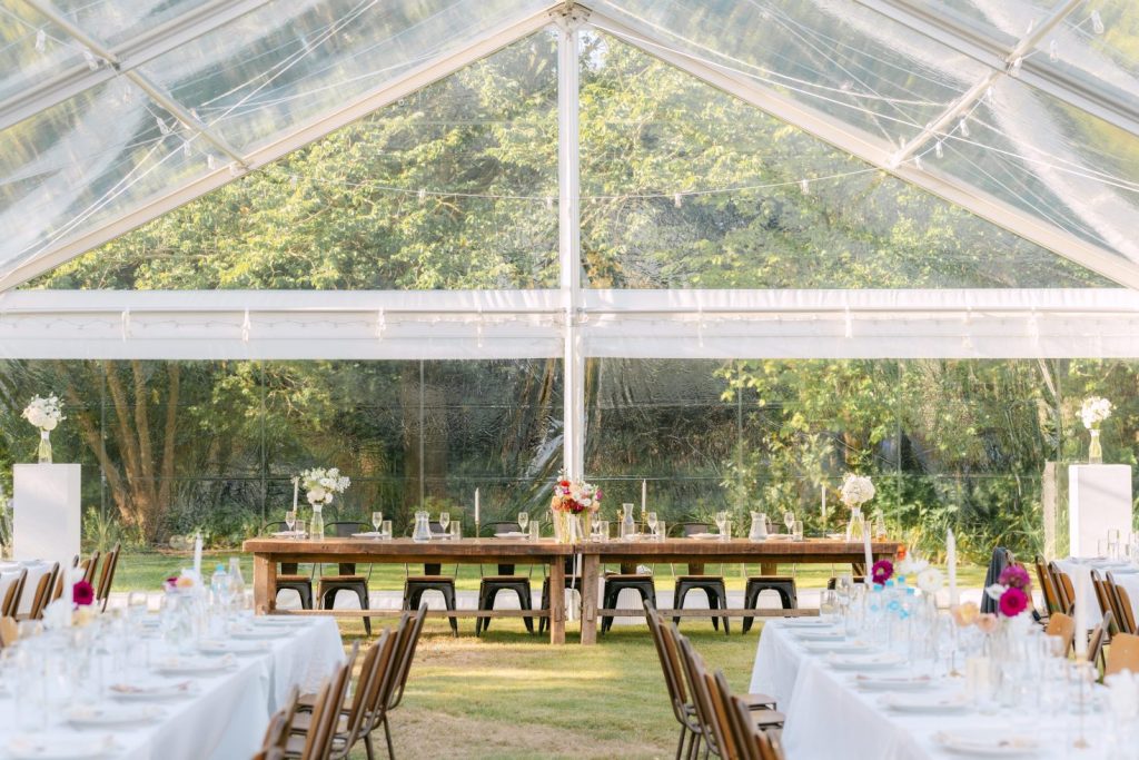 Luxury wedding marquee design: High-angle view looking through a clear glass marquee tent surrounded by lush green trees in Ohoka.