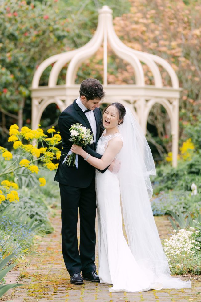 Candid outdoor wedding photography: Bride and groom embracing warmly under a classic garden gazebo.