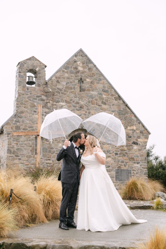 A couple portrait under white umbrellas during a rainy Tekapo day at a Church of the Good Shepherd wedding.